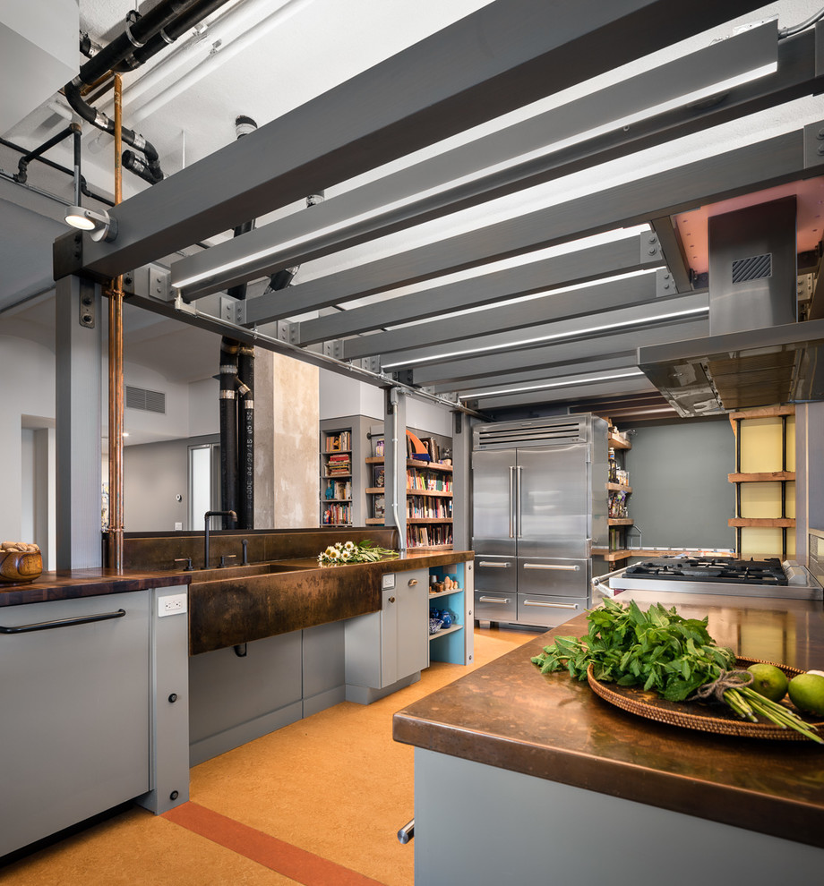Large industrial galley kitchen in New York with an integrated sink, flat-panel cabinets, grey cabinets, copper benchtops, stainless steel appliances, cork floors, brown floor and with island.