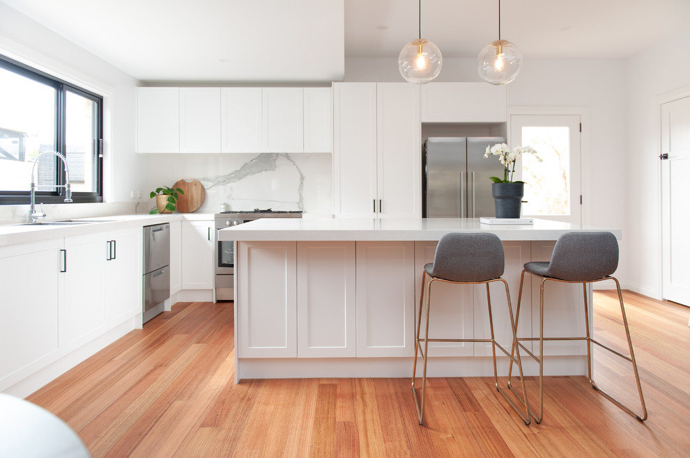Large transitional l-shaped open plan kitchen in Melbourne with an undermount sink, white cabinets, white splashback, stone slab splashback, stainless steel appliances, medium hardwood floors, with island, white benchtop, shaker cabinets and beige floor.