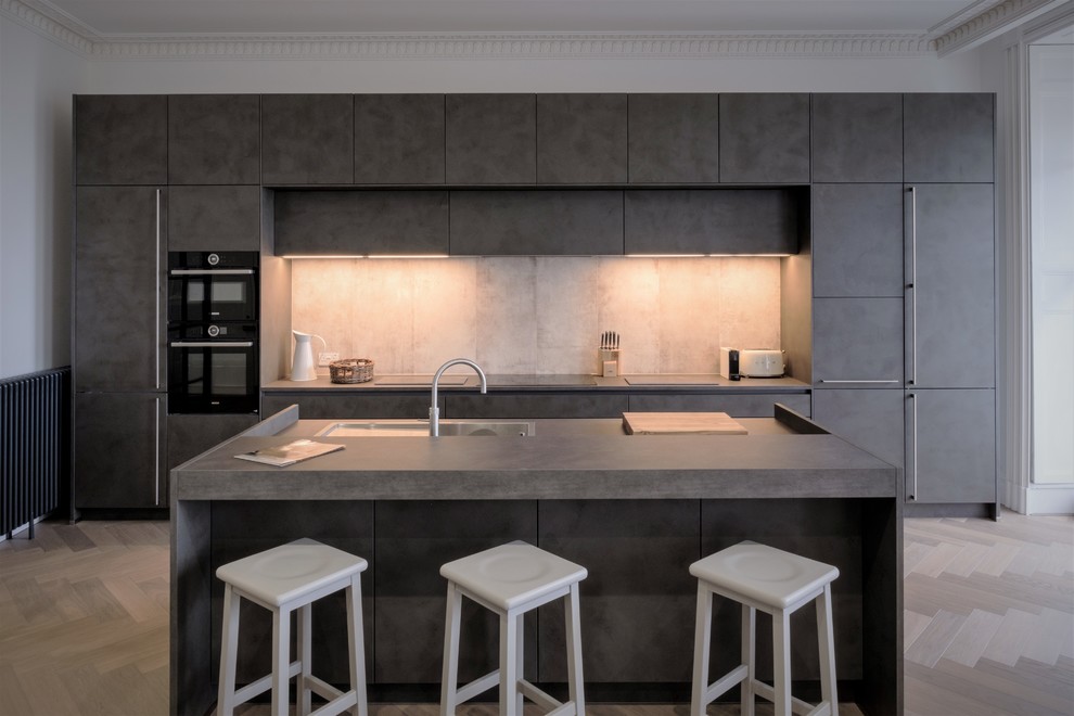 This is an example of a modern kitchen in Other with flat-panel cabinets, grey cabinets, concrete benchtops, white splashback, black appliances, light hardwood floors, with island, beige floor and grey benchtop.
