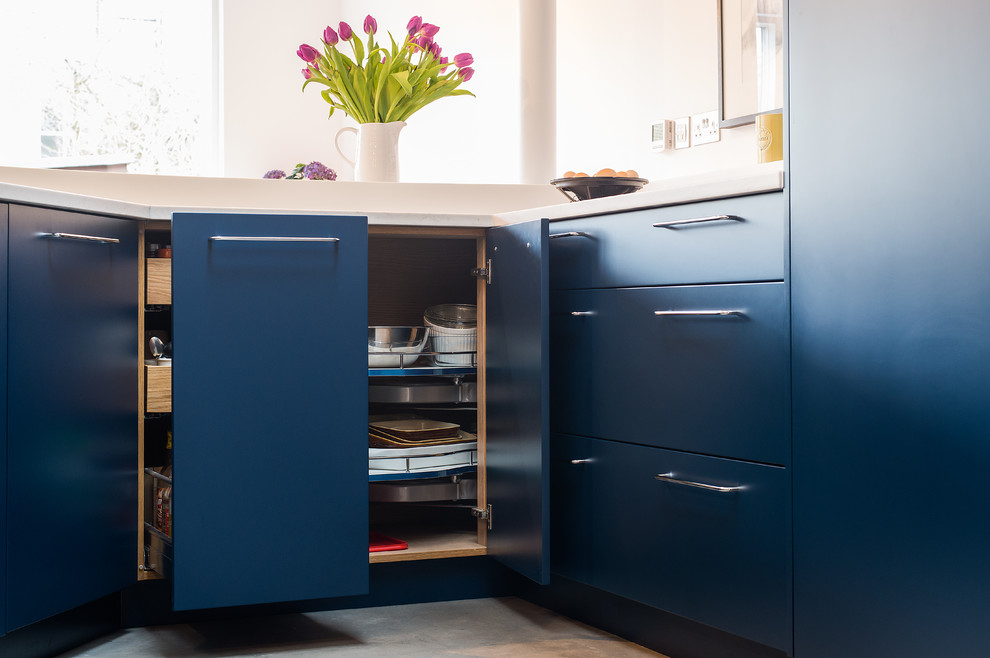 This is an example of a mid-sized eclectic u-shaped open plan kitchen in London with a double-bowl sink, flat-panel cabinets, blue cabinets, solid surface benchtops, panelled appliances, concrete floors and a peninsula.