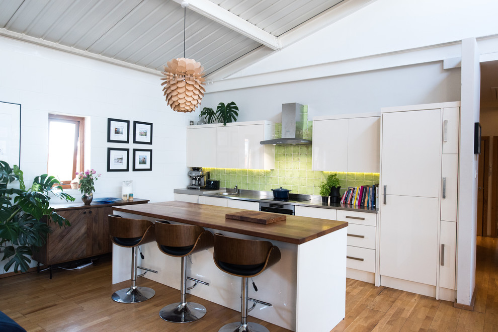 This is an example of a mid-sized transitional single-wall eat-in kitchen in London with an integrated sink, flat-panel cabinets, white cabinets, stainless steel benchtops, green splashback, ceramic splashback, stainless steel appliances, laminate floors, with island, brown floor and grey benchtop.