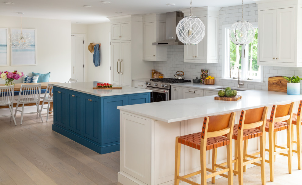 Photo of a beach style l-shaped eat-in kitchen in New York with an undermount sink, recessed-panel cabinets, white cabinets, white splashback, subway tile splashback, stainless steel appliances, light hardwood floors, with island, beige floor and white benchtop.
