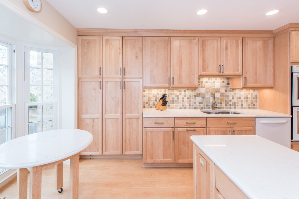Photo of a mid-sized transitional l-shaped eat-in kitchen in DC Metro with a double-bowl sink, shaker cabinets, light wood cabinets, quartzite benchtops, grey splashback, stone tile splashback, stainless steel appliances, vinyl floors, a peninsula and beige floor.