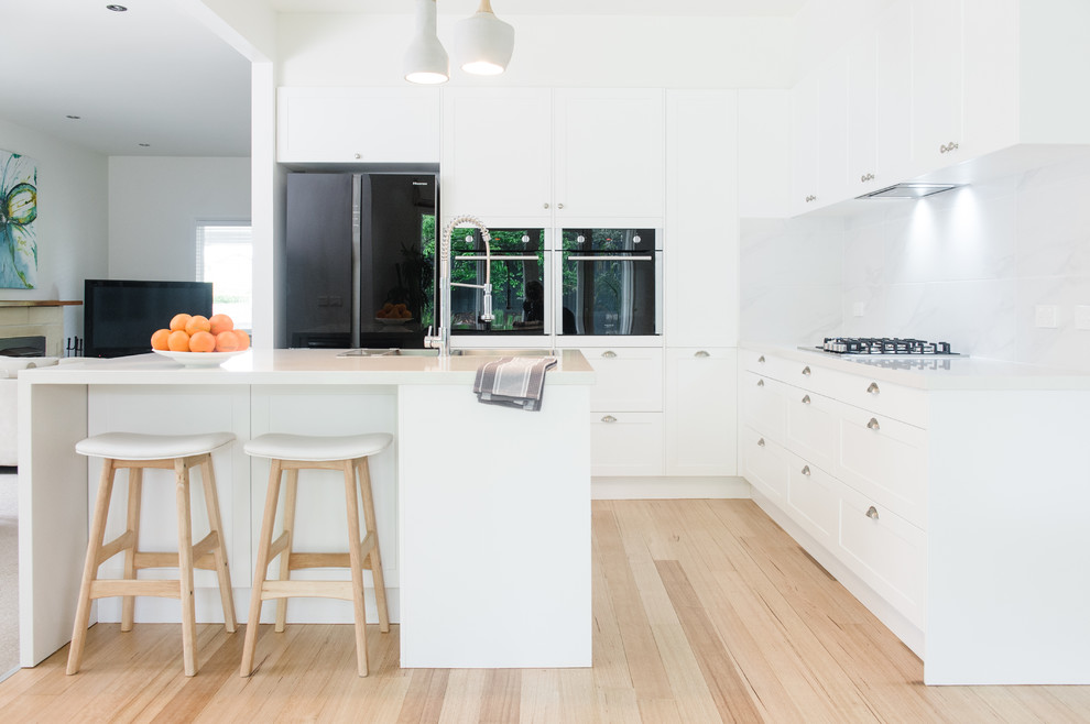 Design ideas for a large scandinavian l-shaped kitchen pantry in Melbourne with a double-bowl sink, shaker cabinets, white cabinets, solid surface benchtops, white splashback, ceramic splashback, black appliances, light hardwood floors and with island.
