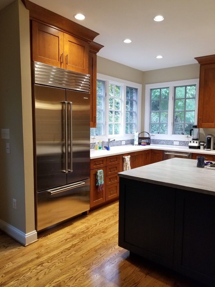 This is an example of a mid-sized transitional l-shaped eat-in kitchen in DC Metro with an integrated sink, recessed-panel cabinets, medium wood cabinets, solid surface benchtops, blue splashback, mosaic tile splashback, stainless steel appliances, medium hardwood floors, a peninsula, brown floor and blue benchtop.