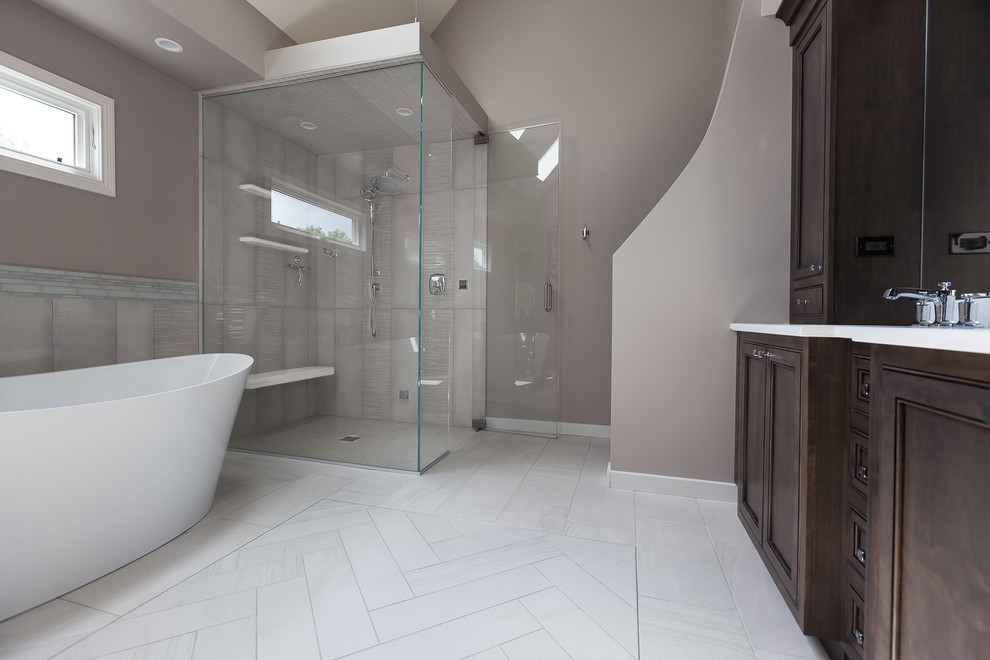 This is an example of a large transitional master bathroom in Chicago with beaded inset cabinets, dark wood cabinets, a freestanding tub, a curbless shower, a two-piece toilet, gray tile, mosaic tile, brown walls, ceramic floors, an undermount sink, engineered quartz benchtops, grey floor, a hinged shower door and grey benchtops.