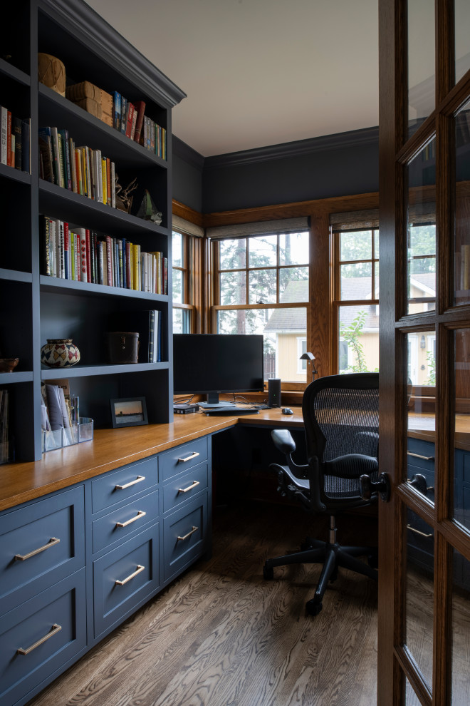Amerikansk inredning av ett mellanstort arbetsrum, med blå väggar, mellanmörkt trägolv, ett inbyggt skrivbord och brunt golv
