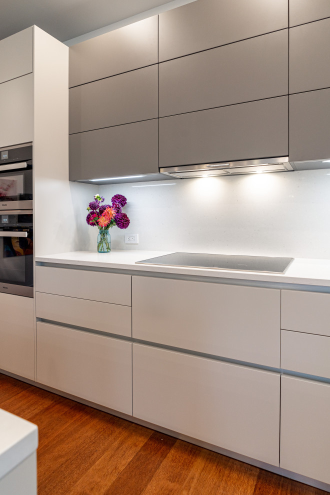 This is an example of a mid-sized modern l-shaped open plan kitchen in Portland with an undermount sink, flat-panel cabinets, beige cabinets, quartz benchtops, white splashback, engineered quartz splashback, panelled appliances, medium hardwood floors, a peninsula, brown floor and white benchtop.