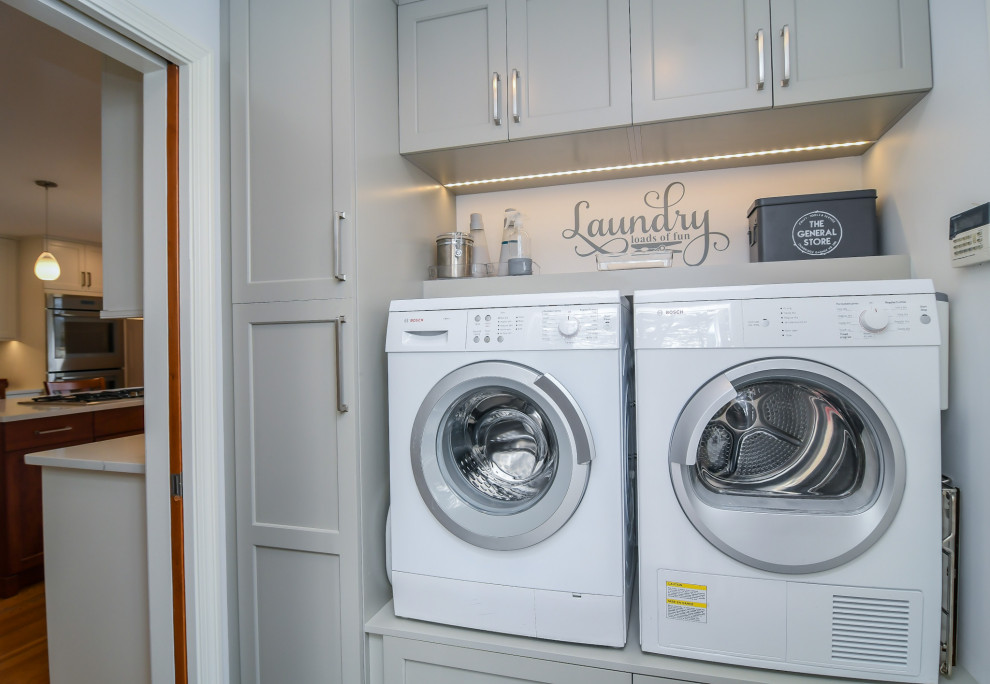 Modelo de lavadero lineal clásico renovado con puertas de armario grises, paredes grises, suelo de baldosas de porcelana, lavadora y secadora juntas y suelo negro