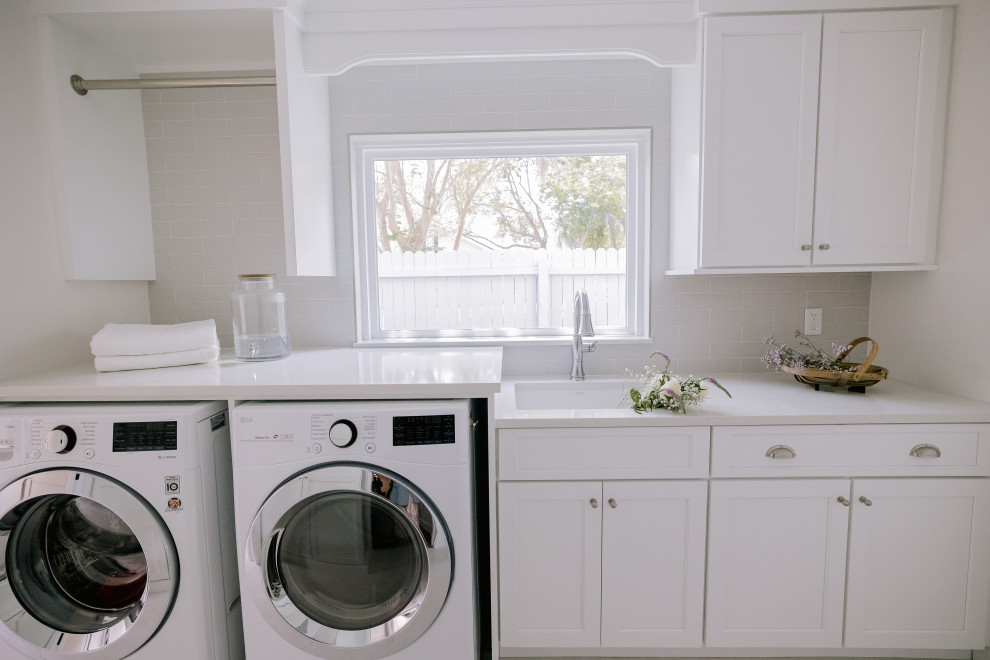 オーランドにある広いトランジショナルスタイルのおしゃれな洗濯室 (I型、ドロップインシンク、インセット扉のキャビネット、白いキャビネット、クオーツストーンカウンター、グレーのキッチンパネル、磁器タイルのキッチンパネル、白い壁、磁器タイルの床、左右配置の洗濯機・乾燥機、白い床、白いキッチンカウンター) の写真