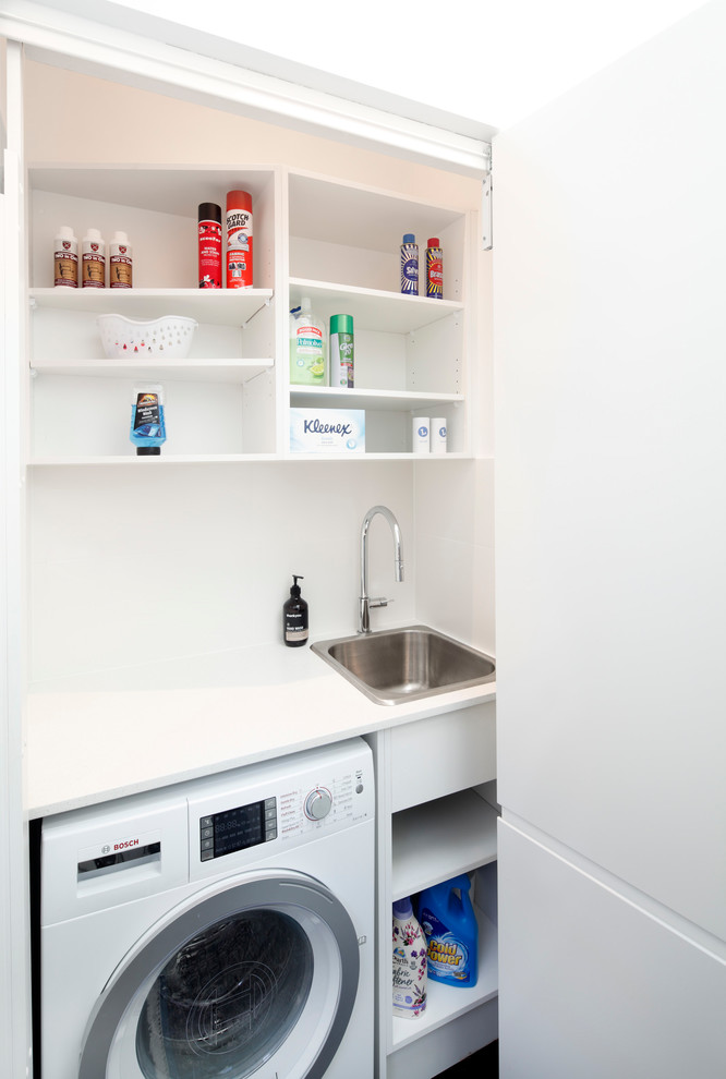 This is an example of a small modern single-wall laundry cupboard in Sydney with a drop-in sink, flat-panel cabinets, white cabinets, quartz benchtops, white walls, dark hardwood floors, an integrated washer and dryer, brown floor and grey benchtop.