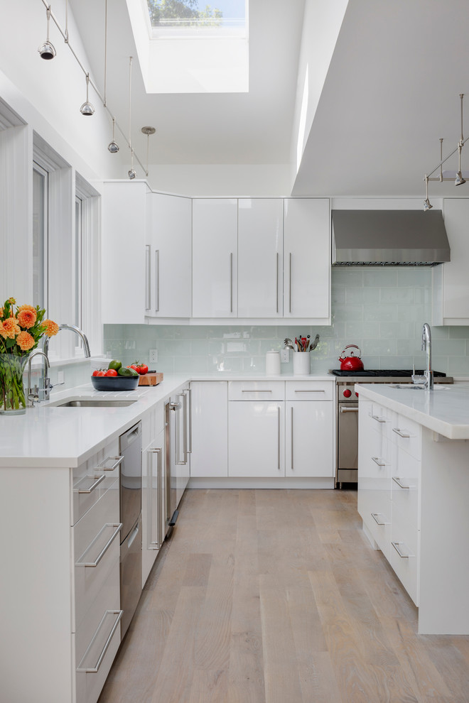 Photo of a mid-sized beach style u-shaped eat-in kitchen in Boston with a drop-in sink, flat-panel cabinets, white cabinets, blue splashback, stainless steel appliances, light hardwood floors, with island, marble benchtops and glass tile splashback.