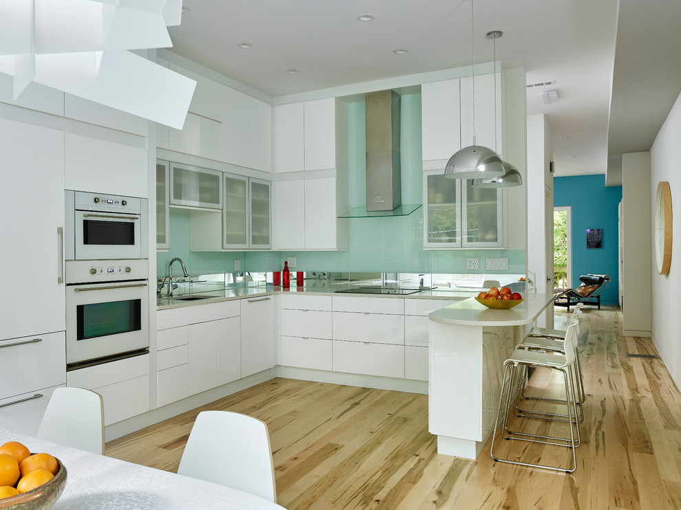 This is an example of a mid-sized modern u-shaped eat-in kitchen in Philadelphia with an undermount sink, flat-panel cabinets, white cabinets, quartz benchtops, white splashback, glass sheet splashback, white appliances and light hardwood floors.