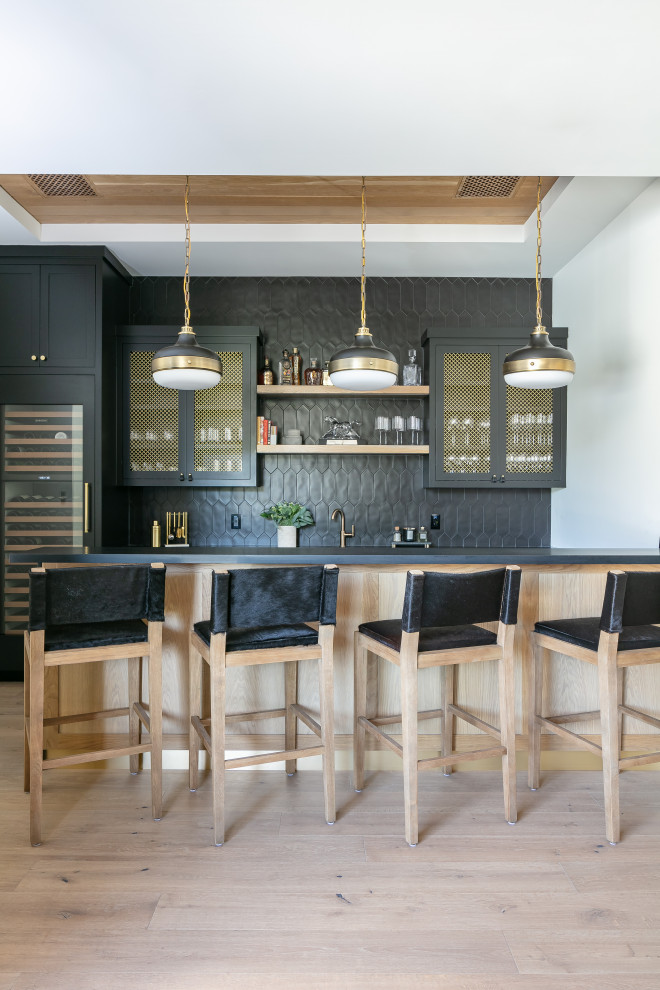 Photo of a country galley seated home bar in Los Angeles with shaker cabinets, black cabinets, black splashback, light hardwood floors, beige floor and black benchtop.