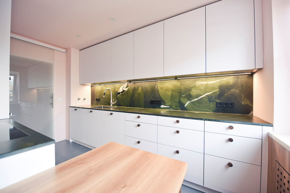 Photo of a mid-sized scandinavian galley separate kitchen in Munich with a single-bowl sink, flat-panel cabinets, white cabinets, granite benchtops, green splashback, stone slab splashback, black appliances, concrete floors, no island, grey floor and green benchtop.