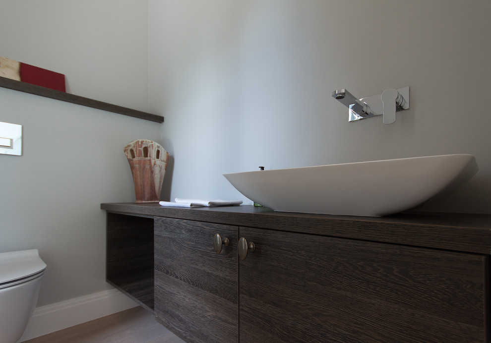 This is an example of a mid-sized modern powder room in London with flat-panel cabinets, dark wood cabinets, a wall-mount toilet, grey walls, medium hardwood floors, a console sink, wood benchtops and brown floor.