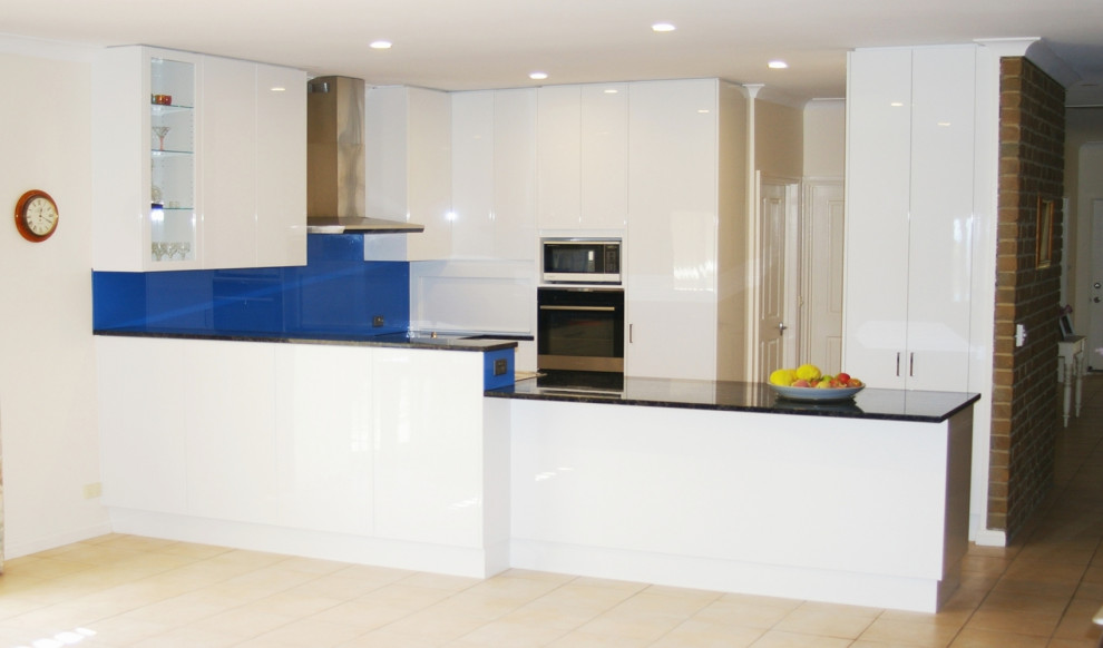 Expansive contemporary u-shaped open plan kitchen with flat-panel cabinets, white cabinets, granite benchtops, blue splashback, glass sheet splashback, stainless steel appliances, ceramic floors, a peninsula, a drop-in sink and beige floor.