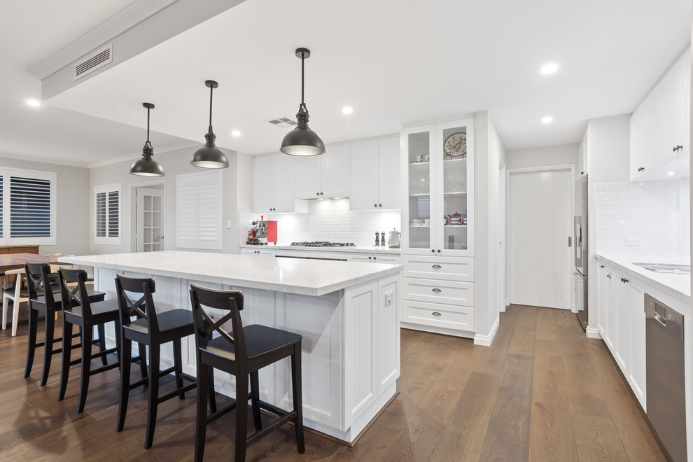 Photo of a beach style galley open plan kitchen in Perth with a double-bowl sink, shaker cabinets, white cabinets, solid surface benchtops, white splashback, subway tile splashback, stainless steel appliances, medium hardwood floors, with island and white benchtop.