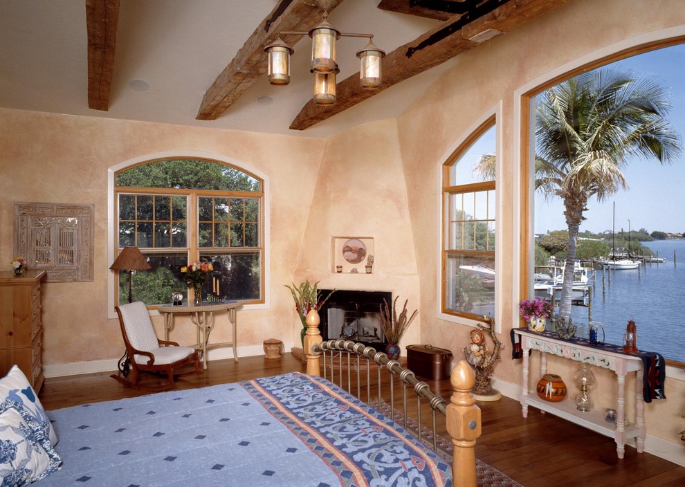 This is an example of a mid-sized mediterranean master bedroom in Tampa with beige walls, medium hardwood floors, a wood stove, a plaster fireplace surround and brown floor.