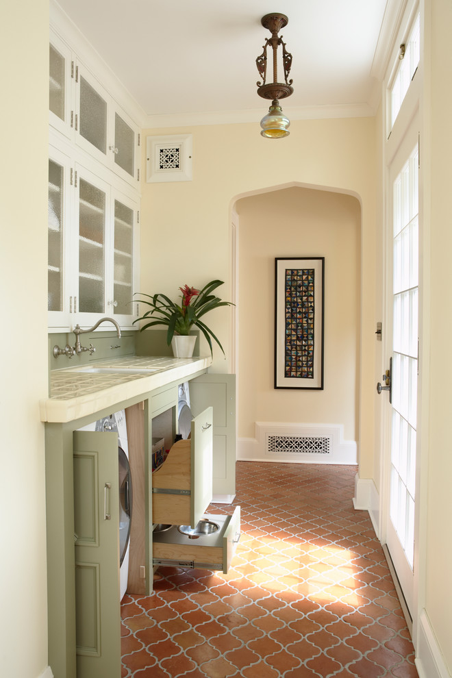 Design ideas for a traditional single-wall utility room in Minneapolis with an undermount sink, recessed-panel cabinets, green cabinets, tile benchtops, terra-cotta floors, a concealed washer and dryer and beige walls.