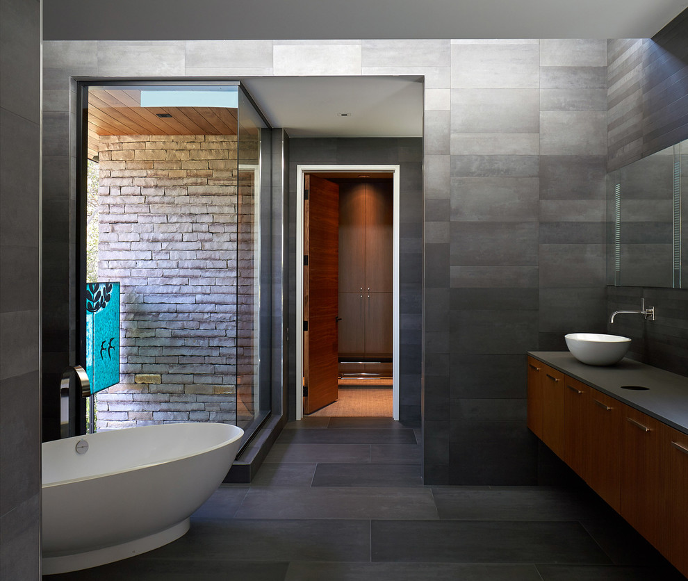 Photo of a mid-sized modern master bathroom in Austin with a freestanding tub, an open shower, gray tile, flat-panel cabinets, medium wood cabinets, black walls, a vessel sink, black floor and black benchtops.