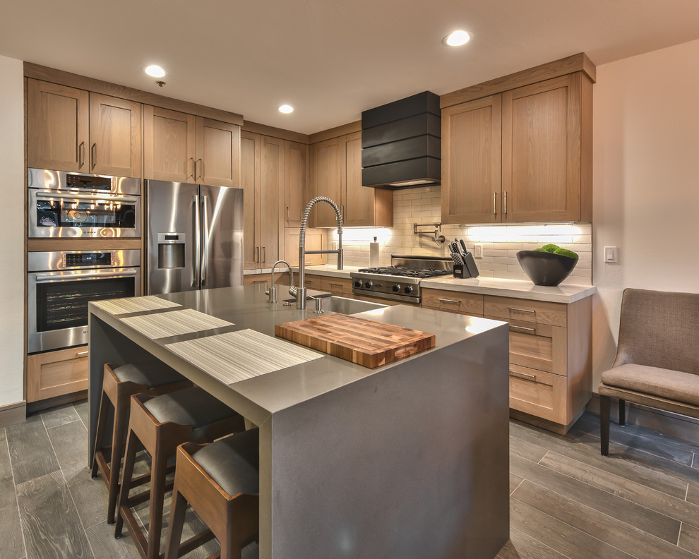 This is an example of a country l-shaped kitchen in Salt Lake City with a farmhouse sink, shaker cabinets, medium wood cabinets, beige splashback, subway tile splashback, stainless steel appliances, medium hardwood floors, with island, grey floor and grey benchtop.