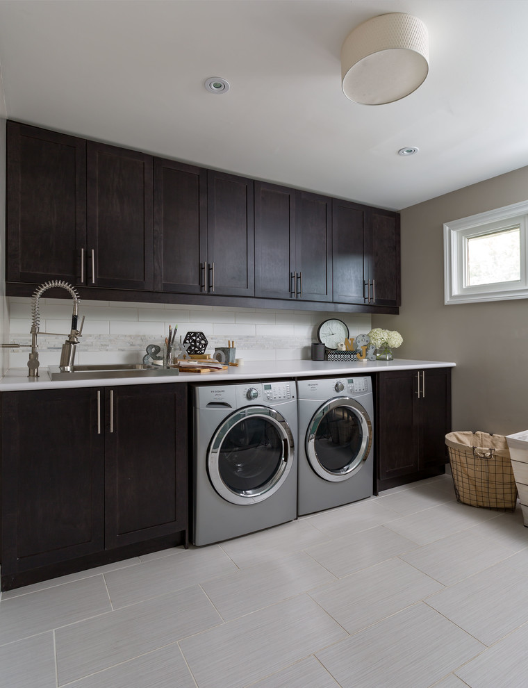 Large transitional l-shaped dedicated laundry room in Toronto with an undermount sink, shaker cabinets, grey splashback, subway tile splashback, porcelain floors, grey floor, quartz benchtops, beige walls, a side-by-side washer and dryer, white benchtop and dark wood cabinets.