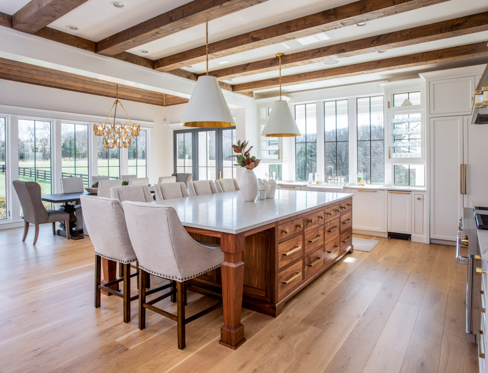 This is an example of a large country l-shaped open plan kitchen in Nashville with a farmhouse sink, beaded inset cabinets, medium wood cabinets, marble benchtops, white splashback, stainless steel appliances, light hardwood floors, with island, brown floor, white benchtop and exposed beam.