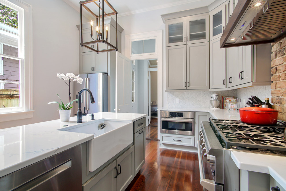 This is an example of a mid-sized traditional l-shaped separate kitchen in New Orleans with a farmhouse sink, recessed-panel cabinets, grey cabinets, quartzite benchtops, white splashback, marble splashback, stainless steel appliances, medium hardwood floors, with island, brown floor and white benchtop.