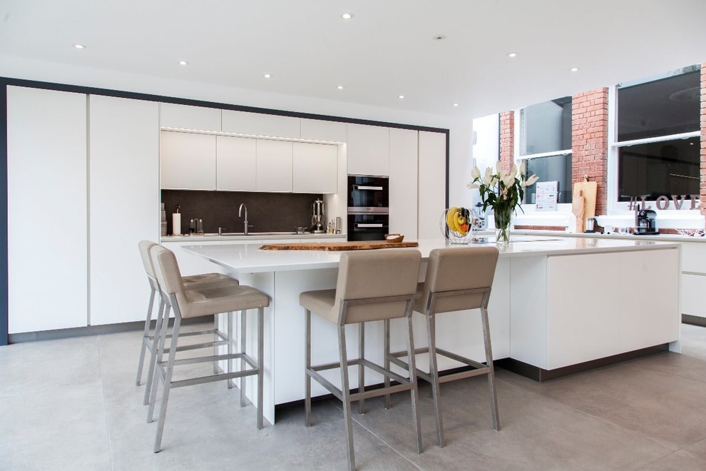 Design ideas for a large contemporary l-shaped kitchen in Hampshire with a triple-bowl sink, flat-panel cabinets, white cabinets, grey splashback, stone tile splashback, black appliances, ceramic floors, with island and grey floor.