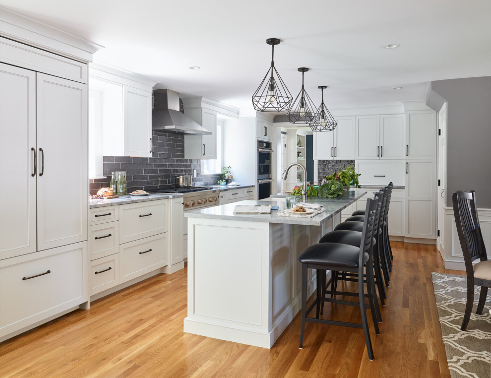 Photo of a transitional l-shaped kitchen in Boston with a farmhouse sink, recessed-panel cabinets, white cabinets, grey splashback, stainless steel appliances, medium hardwood floors, with island, brown floor and grey benchtop.