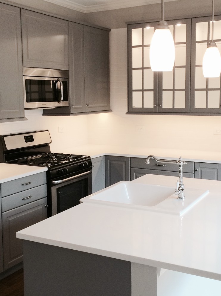 This is an example of a mid-sized modern l-shaped separate kitchen in Chicago with a farmhouse sink, raised-panel cabinets, grey cabinets, solid surface benchtops, black appliances, dark hardwood floors, with island, brown floor, white splashback and subway tile splashback.