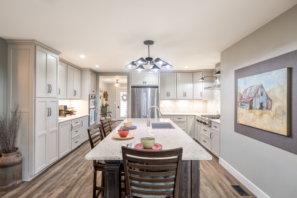 This is an example of a transitional u-shaped kitchen in Other with a double-bowl sink, shaker cabinets, white cabinets, white splashback, stainless steel appliances, medium hardwood floors, with island, brown floor and white benchtop.