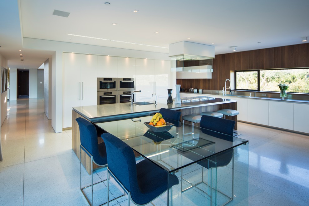 Large contemporary l-shaped open plan kitchen in Orange County with with island, an undermount sink, flat-panel cabinets, white cabinets, stainless steel benchtops, metallic splashback, metal splashback, stainless steel appliances, porcelain floors and white floor.