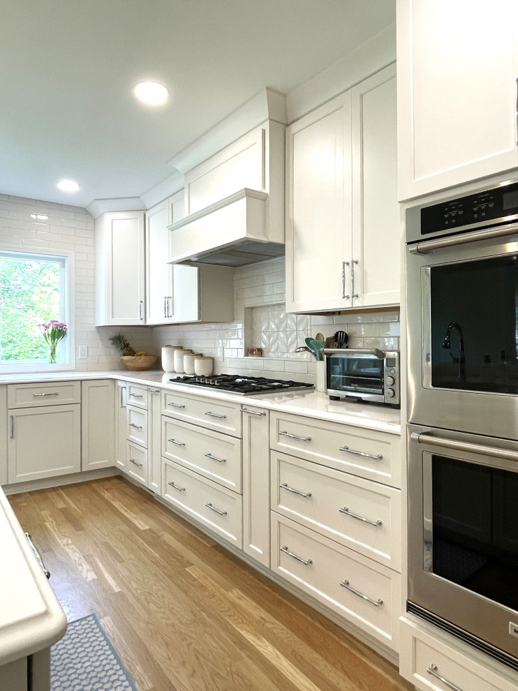 This is an example of a large classic l-shaped kitchen/diner in Detroit with a submerged sink, shaker cabinets, white cabinets, engineered stone countertops, white splashback, ceramic splashback, stainless steel appliances, medium hardwood flooring, an island, brown floors and white worktops.