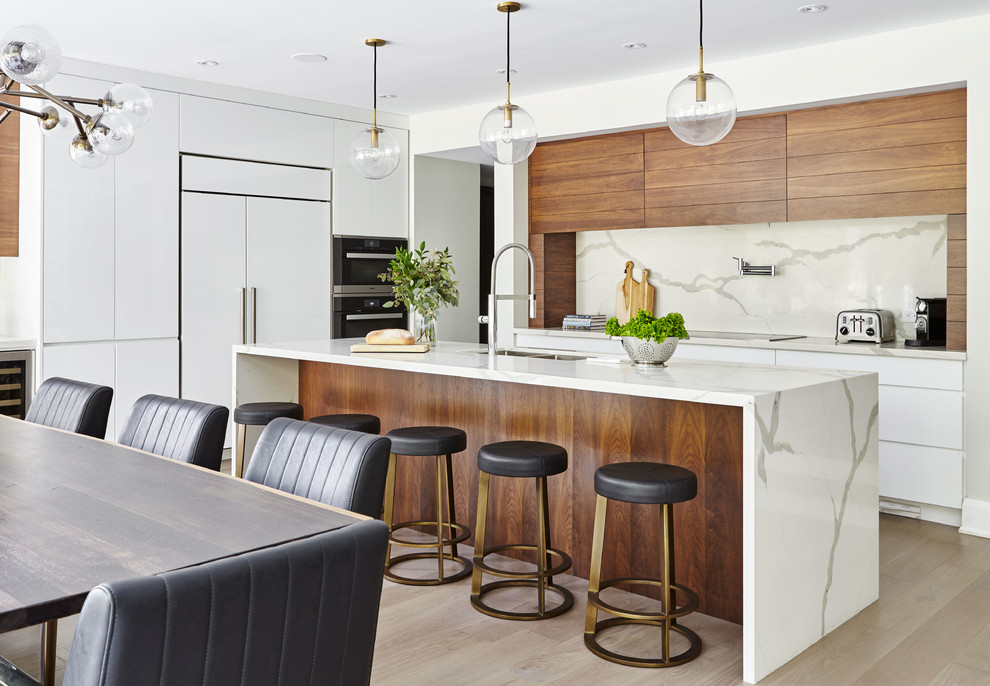 Modern open plan kitchen in Toronto with a drop-in sink, flat-panel cabinets, white cabinets, quartzite benchtops, white splashback, stone slab splashback, stainless steel appliances, light hardwood floors, with island and brown floor.