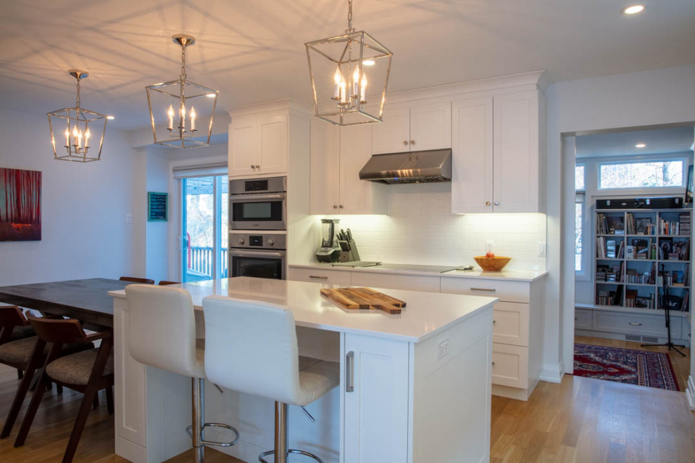 Eat-in kitchen - large transitional l-shaped light wood floor eat-in kitchen idea in Ottawa with a farmhouse sink, shaker cabinets, white cabinets, quartz countertops, white backsplash, porcelain backsplash, stainless steel appliances, an island and white countertops