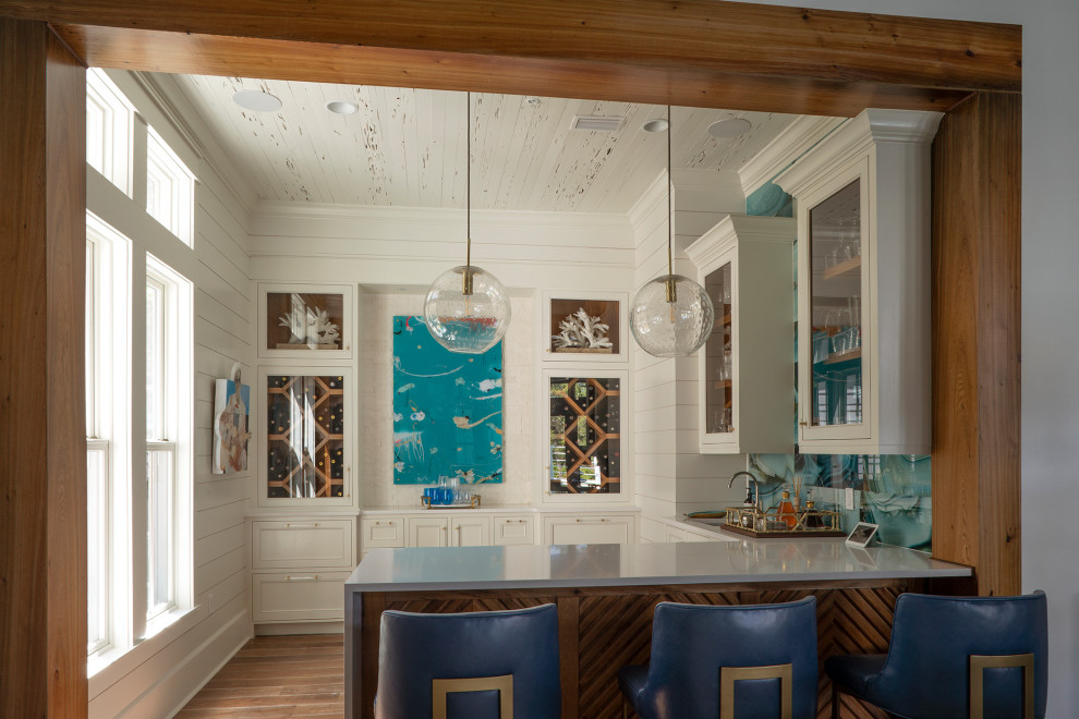 Inspiration for a mid-sized beach style u-shaped seated home bar in Other with glass-front cabinets, white cabinets, quartzite benchtops, blue splashback, medium hardwood floors, brown floor and white benchtop.