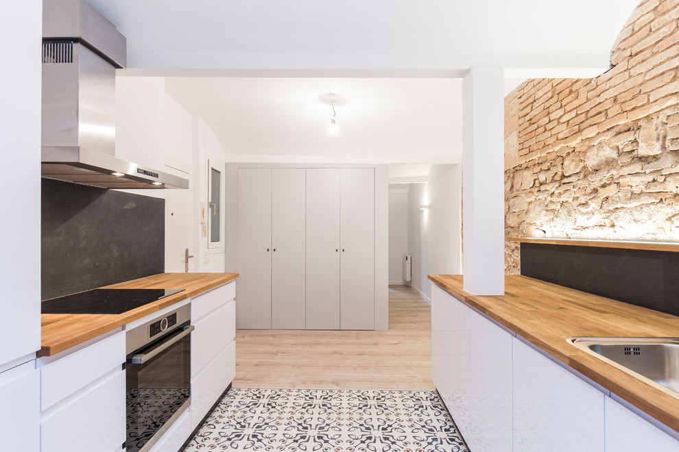 This is an example of a small mediterranean single-wall open plan kitchen in Barcelona with a farmhouse sink, recessed-panel cabinets, white cabinets, wood benchtops, black splashback, slate splashback, stainless steel appliances, cement tiles, no island and multi-coloured floor.