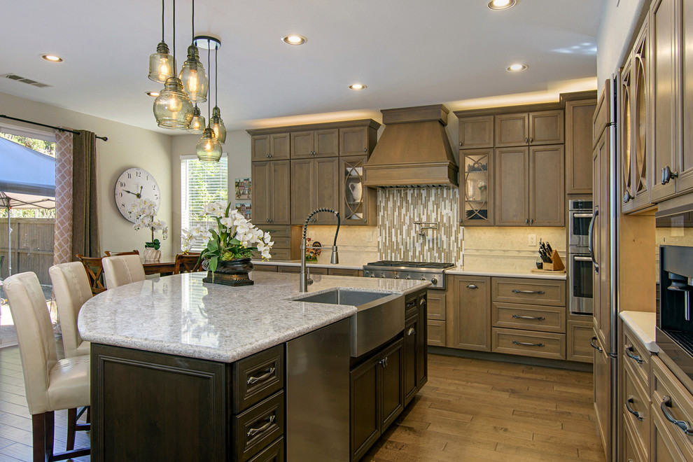 Example of a large transitional l-shaped medium tone wood floor and brown floor open concept kitchen design in San Diego with a farmhouse sink, recessed-panel cabinets, medium tone wood cabinets, beige backsplash, paneled appliances, an island, quartz countertops and travertine backsplash