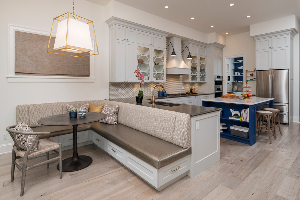 This is an example of a beach style eat-in kitchen in Vancouver with a double-bowl sink, glass-front cabinets, white cabinets, white splashback, stainless steel appliances, light hardwood floors, with island, beige floor and black benchtop.