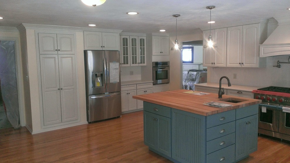 Example of a transitional l-shaped medium tone wood floor kitchen design with a farmhouse sink, raised-panel cabinets, white cabinets, white backsplash, subway tile backsplash, stainless steel appliances and an island