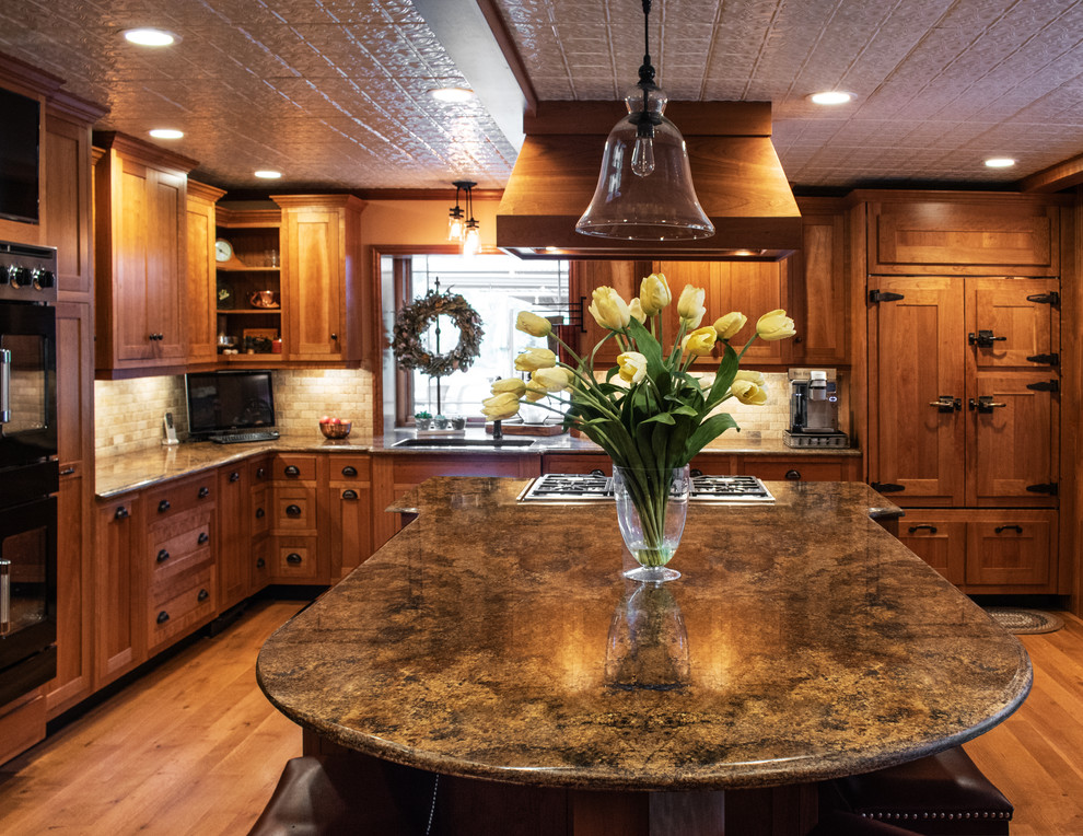 Photo of a country l-shaped eat-in kitchen in New York with an undermount sink, shaker cabinets, medium wood cabinets, granite benchtops, beige splashback, limestone splashback, black appliances, medium hardwood floors, with island, brown floor and multi-coloured benchtop.