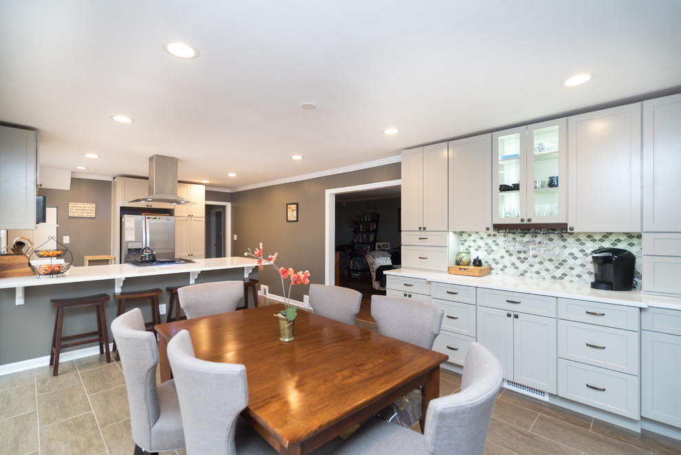 Photo of a mid-sized transitional l-shaped open plan kitchen in DC Metro with a farmhouse sink, shaker cabinets, white cabinets, quartz benchtops, grey splashback, porcelain splashback, stainless steel appliances, laminate floors and a peninsula.