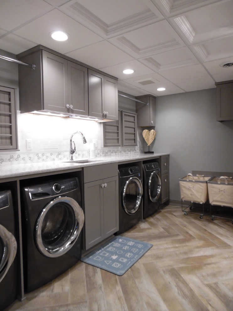 Photo of a large traditional utility room in Milwaukee with an undermount sink, recessed-panel cabinets, grey cabinets, quartz benchtops, vinyl floors, a side-by-side washer and dryer, brown floor and white benchtop.