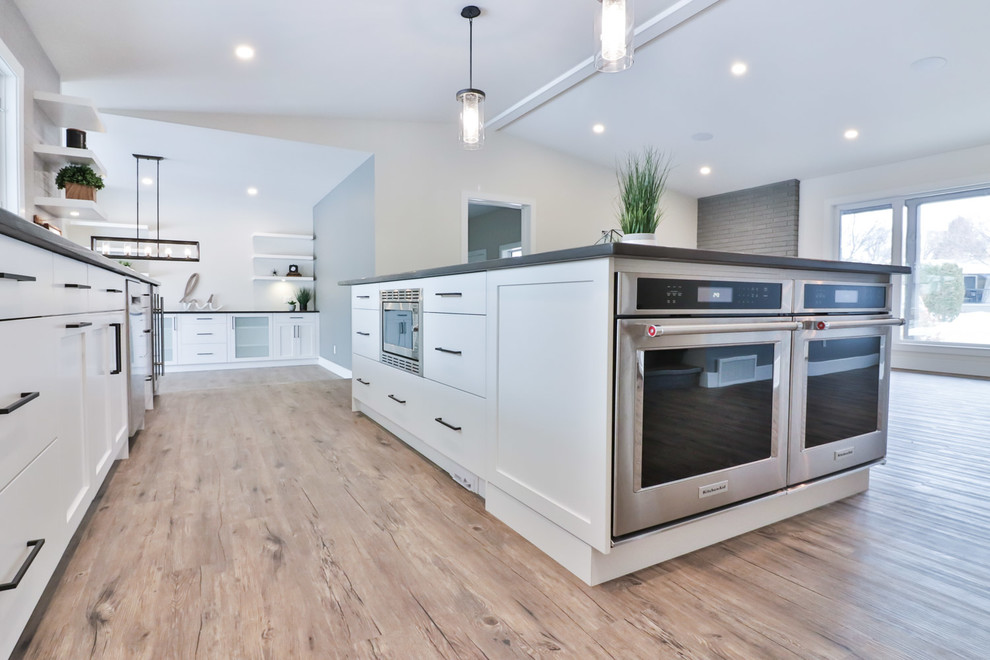 Large transitional l-shaped vinyl floor and brown floor open concept kitchen photo with an undermount sink, flat-panel cabinets, white cabinets, quartz countertops, gray backsplash, porcelain backsplash, stainless steel appliances and an island