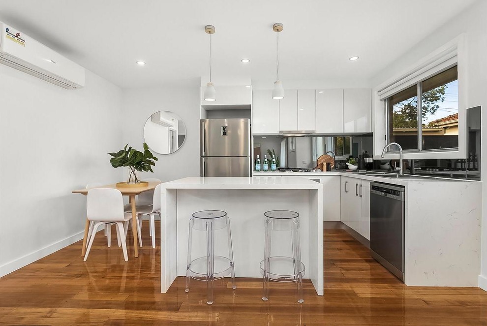 This is an example of a mid-sized modern l-shaped kitchen pantry in Melbourne with a double-bowl sink, open cabinets, white cabinets, solid surface benchtops, glass sheet splashback, stainless steel appliances, medium hardwood floors, with island and white benchtop.