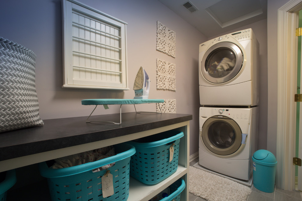 This is an example of a mid-sized traditional galley dedicated laundry room in Indianapolis with open cabinets, solid surface benchtops, purple walls, vinyl floors, a stacked washer and dryer and grey floor.
