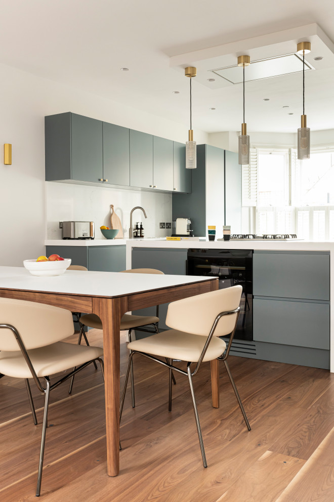 This is an example of a mid-sized contemporary u-shaped open plan kitchen in London with an integrated sink, flat-panel cabinets, blue cabinets, solid surface benchtops, white splashback, engineered quartz splashback, stainless steel appliances, medium hardwood floors, with island, brown floor and white benchtop.