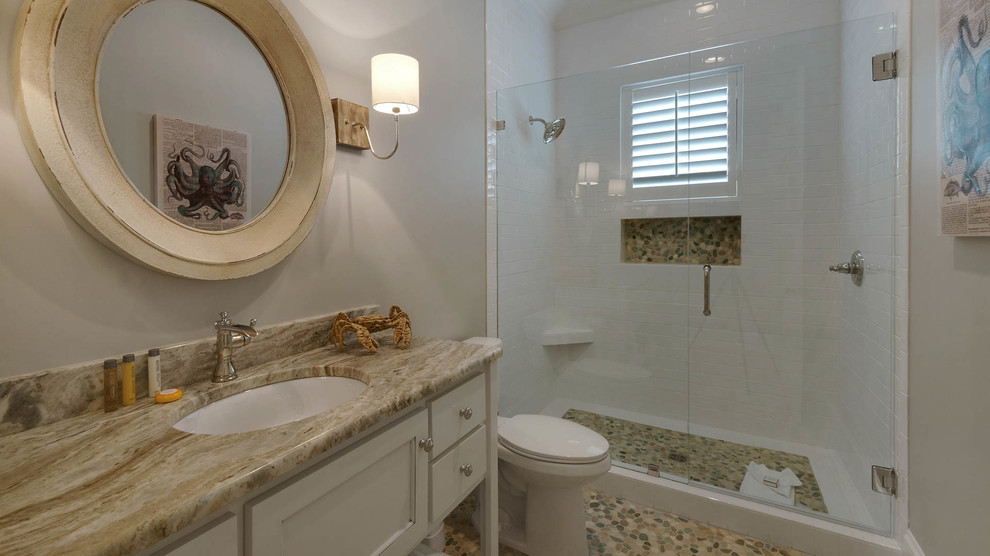This is an example of a mid-sized beach style 3/4 bathroom in Miami with recessed-panel cabinets, white cabinets, an alcove shower, a two-piece toilet, beige tile, stone slab, grey walls, pebble tile floors, an undermount sink and granite benchtops.